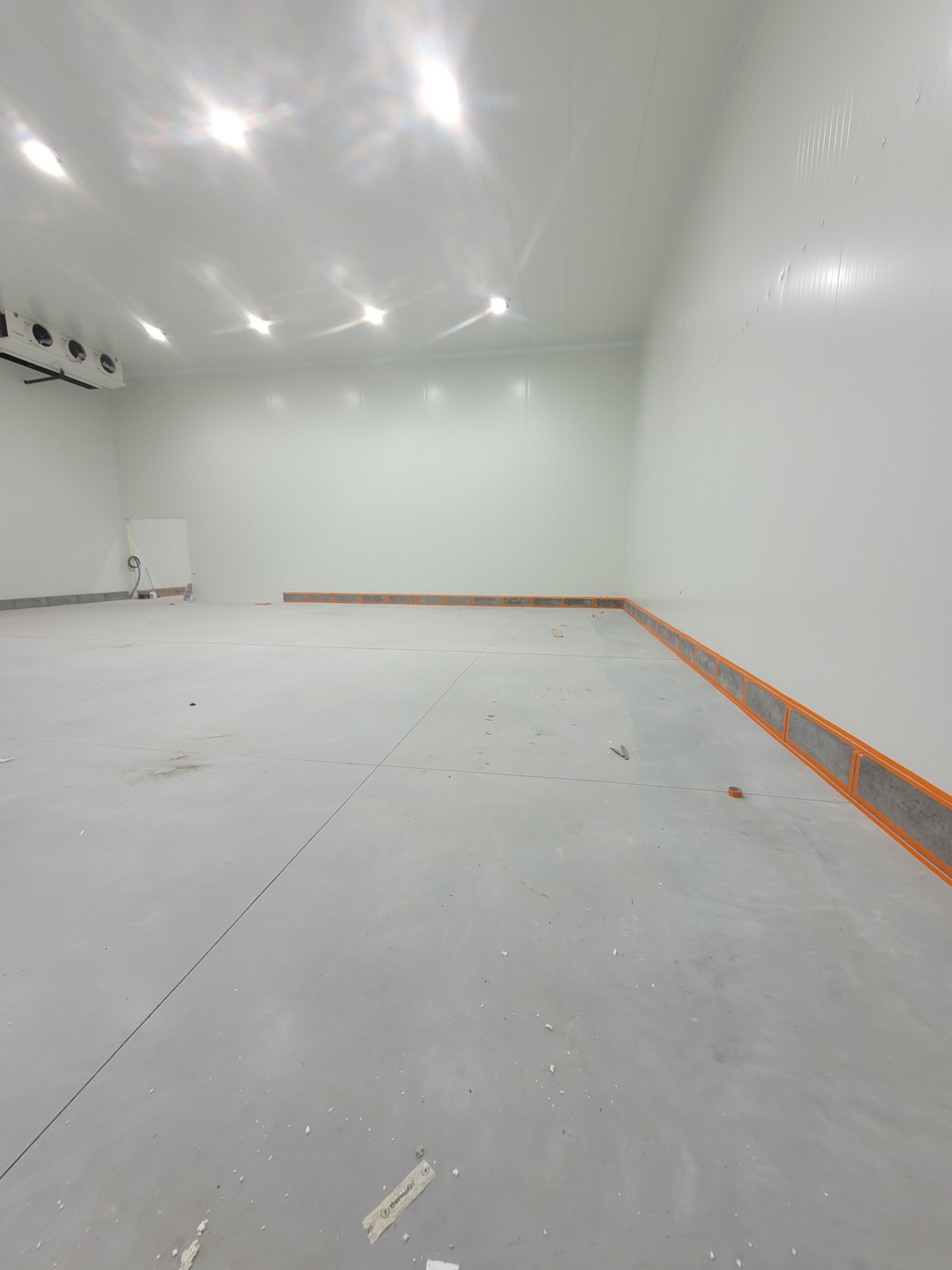 Wnętrze chłodni z panelami warstwowymi i listwami przypodłogowymi. Widoczne elementy instalacji chłodniczej na suficie. Jasne oświetlenie.