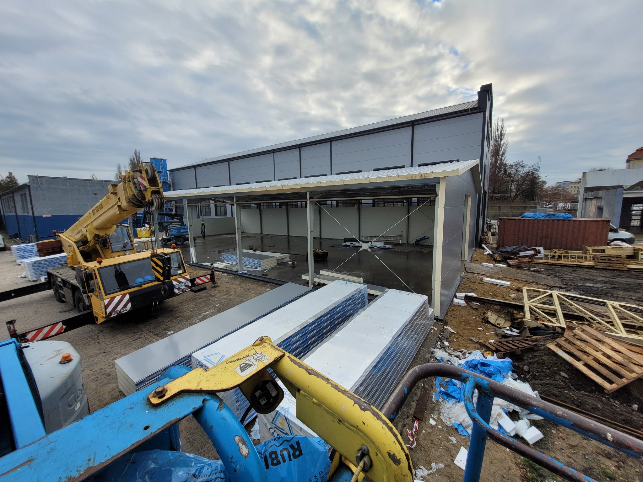 Montaż hali z płyt warstwowych w trakcie budowy. Widoczny dźwig, ułożone płyty i częściowo zmontowana konstrukcja. Pochmurne niebo w tle.