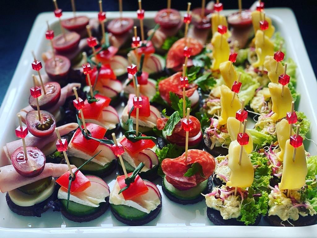 Elegancka taca z różnorodnymi, apetycznymi kanapeczkami na przyjęcie, udekorowanymi czerwonymi kostkami na wykałaczkach. Widoczne kompozycje z wędlin, serów i warzyw.