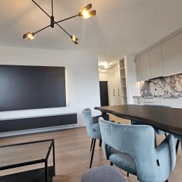 Paweł Gębura - Jasne wnętrze z nowoczesną kuchnią, marmurowym blatem i oświetleniem punktowym, widoczne krzesła z szarego weluru przy ciemnym drewnianym stole, minimalistyczny czarny panel na ścianie.