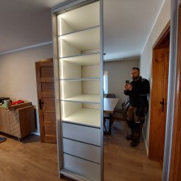 Paweł Gębura - Wnętrze pokoju z nową szafą wnękową z przesuwnymi drzwiami lustrzanymi i oświetleniem LED na półkach, widoczne odbicie mężczyzny robiącego zdjęcie telefonem.