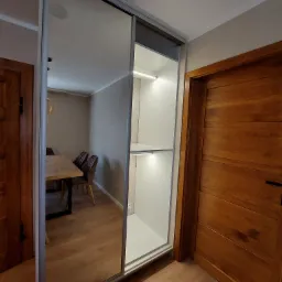 Nowoczesna szafa wnękowa z przesuwnymi drzwiami lustrzanymi i oświetleniem LED, widok z boku na tle drewnianych drzwi i fragmentu jadalni.