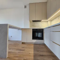 Nowoczesna kuchnia z beżowymi szafkami górnymi i białymi dolnymi, oświetleniem LED podszafkowym i blatem imitującym marmur, widziana z niskiego kąta.