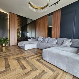 Paweł Gębura - Nowoczesny salon z szarą, modułową sofą narożną, parkietem w jodełkę i ścianami z drewnianymi panelami. Widoczny żyrandol i roślina w złotej doniczce.