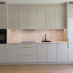 Paweł Gębura - Jasnoszara zabudowa kuchenna z marmurowym blatem i złotymi uchwytami, oświetlona listwą LED. Nowoczesny design z czarną armaturą i sprzętem AGD.