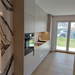 Paweł Gębura - Jasna kuchnia na wymiar z czarną armaturą i piekarnikiem, marmurowy panel boczny, parkiet w jodełkę, okno balkonowe z widokiem na zieleń.