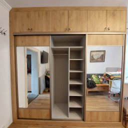 Paweł Gębura - Drewniana szafa wnękowa z przesuwnymi drzwiami lustrzanymi, widok na wnętrze z półkami. Nadstawka z szafkami. Widoczne odbicie pokoju w lustrach.