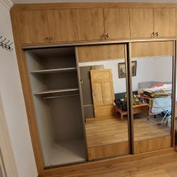 Paweł Gębura - Jasna szafa przesuwna z lustrami, drewnopodobne wykończenie, widoczne półki i drążek na ubrania. W lustrze odbicie pokoju z meblami i oknem.