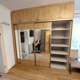 Paweł Gębura - Jasna szafa wnękowa z przesuwnymi drzwiami lustrzanymi, z górnymi szafkami, dopasowana do wnętrza pokoju. Widoczne półki i krzesło obok.