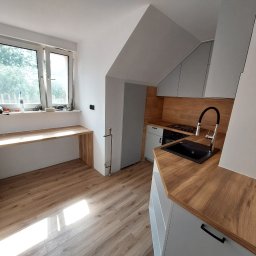 Paweł Gębura - Jasna kuchnia w skosach z drewnianym blatem i podłogą. Okno z blatem roboczym. Szare szafki i czarny zlew. Ujęcie z perspektywy podłogi, widok na całe pomieszczenie.