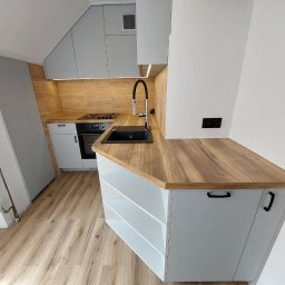 Paweł Gębura - Szara kuchnia z drewnianym blatem i płytkami imitującymi drewno na ścianie. Uwagę przykuwa czarny zlew i bateria. Widoczna płyta grzewcza i piekarnik.