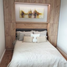 Paweł Gębura - Drewniana zabudowa łóżka z szafkami i półkami, oświetlenie LED, obraz z motywem żaglówek. Jasna pościel i poduszki. Aranżacja sypialni w neutralnych barwach.