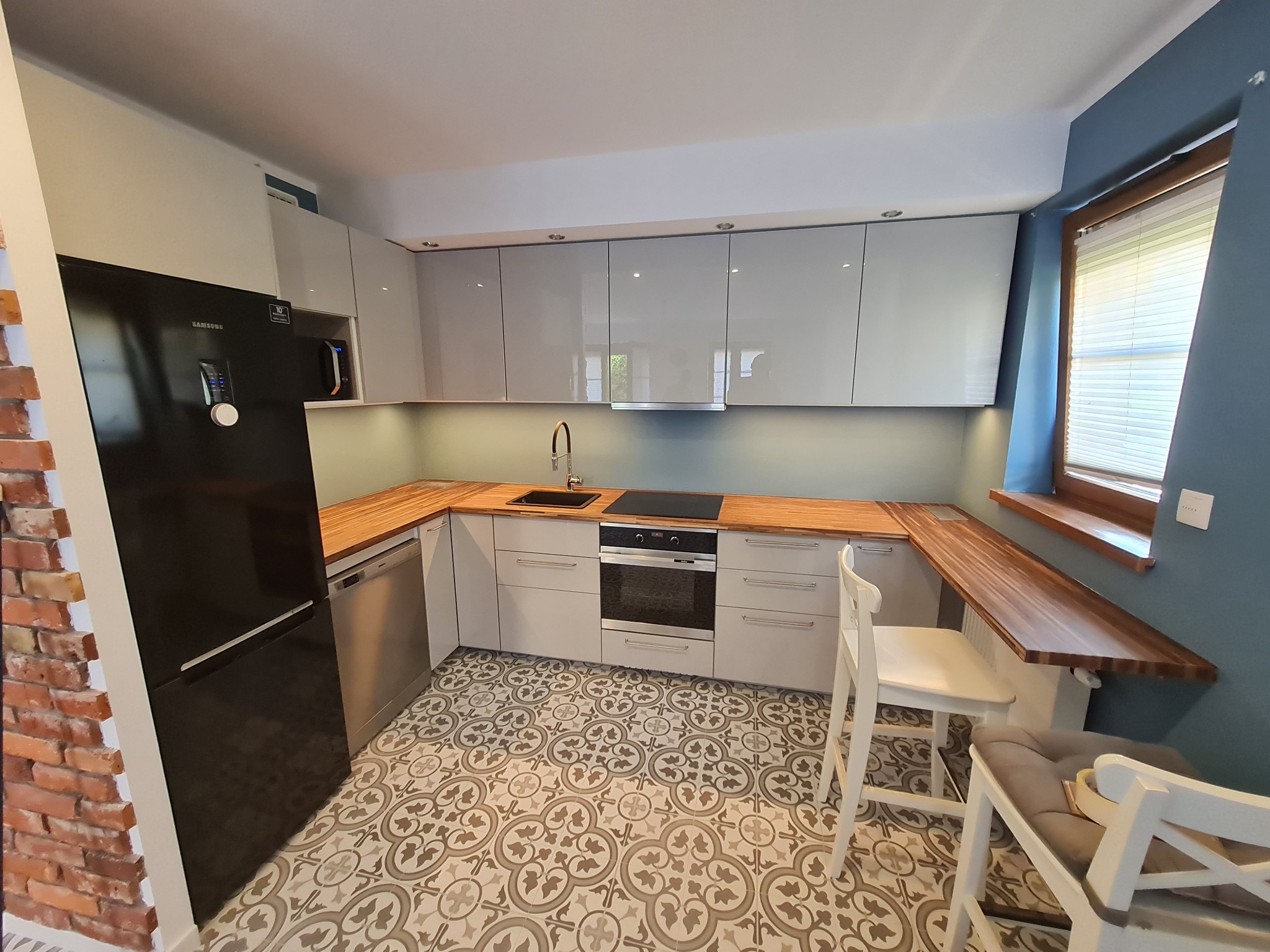 Jasnoszara kuchnia z drewnianymi blatami, czarną lodówką Samsung i wzorzystą podłogą, widok z perspektywy podłogi na ścianę z cegły.