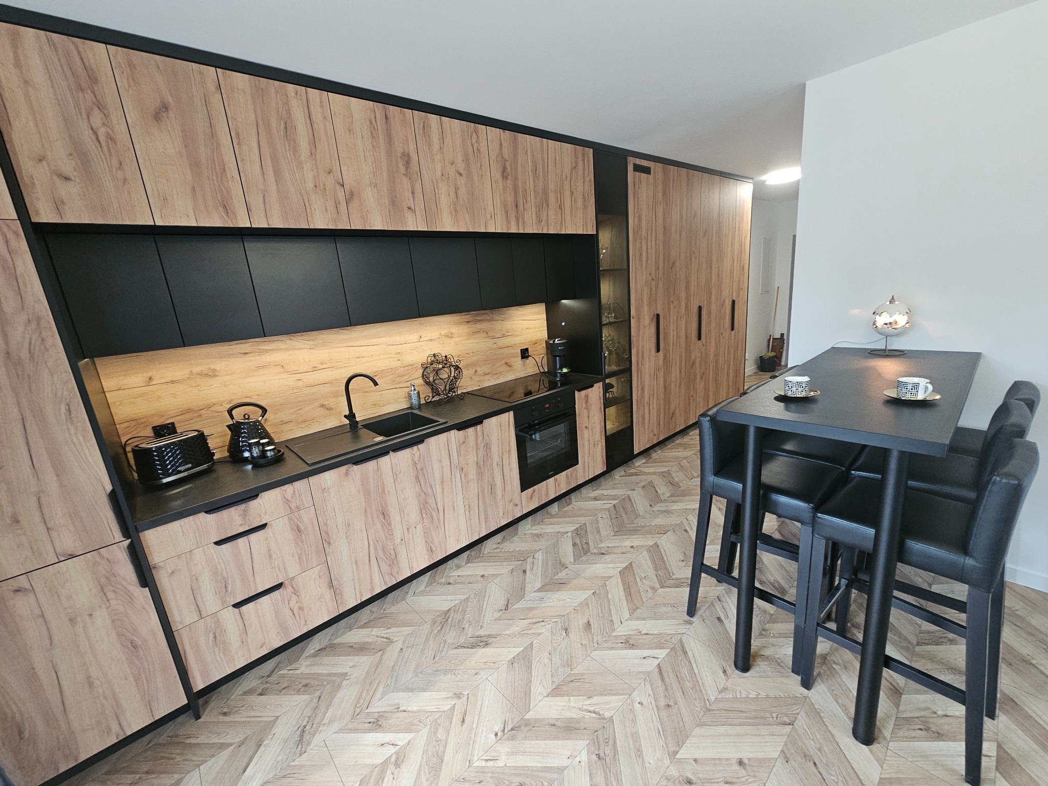 Nowoczesna kuchnia z drewnianymi frontami i czarnymi akcentami, oświetlona ciepłym światłem, z blatem z czarnego kamienia i podłogą w jodełkę, uzupełniona wysokim stołem z krzesłami barowymi.