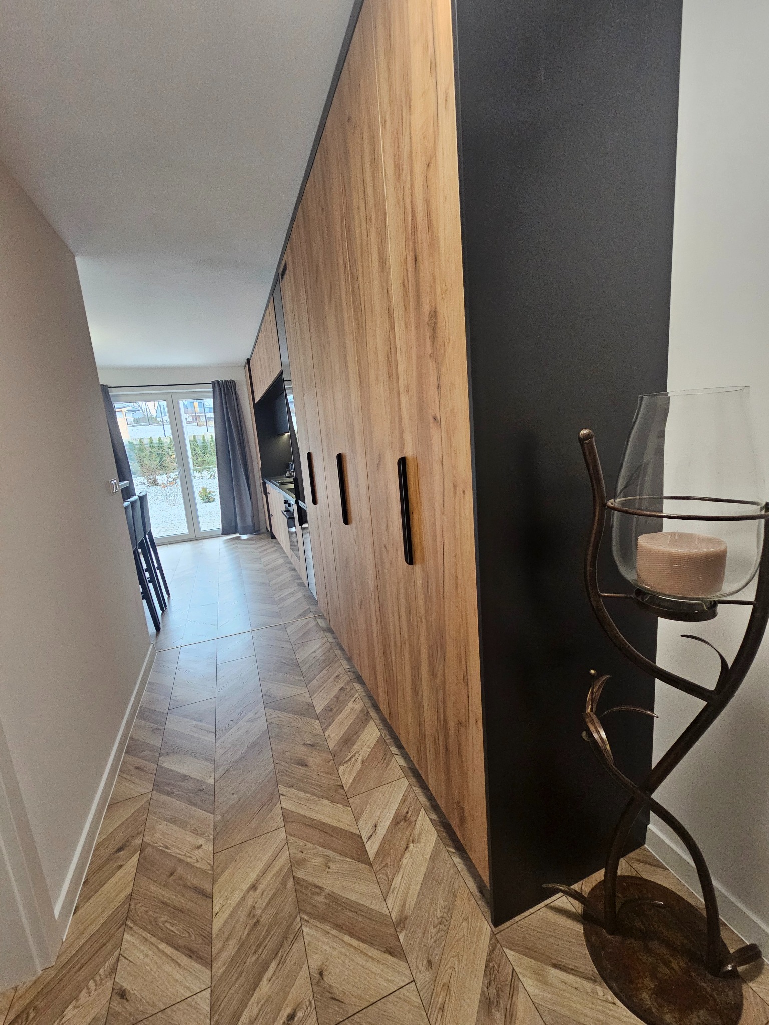 Długi korytarz z podłogą w jodełkę i zabudową meblową z frontami w kolorze drewna, zakończoną czarnym segmentem, z widokiem na kuchnię i ogród w tle oraz dekoracyjnym świecznikiem w pierwszym planie.