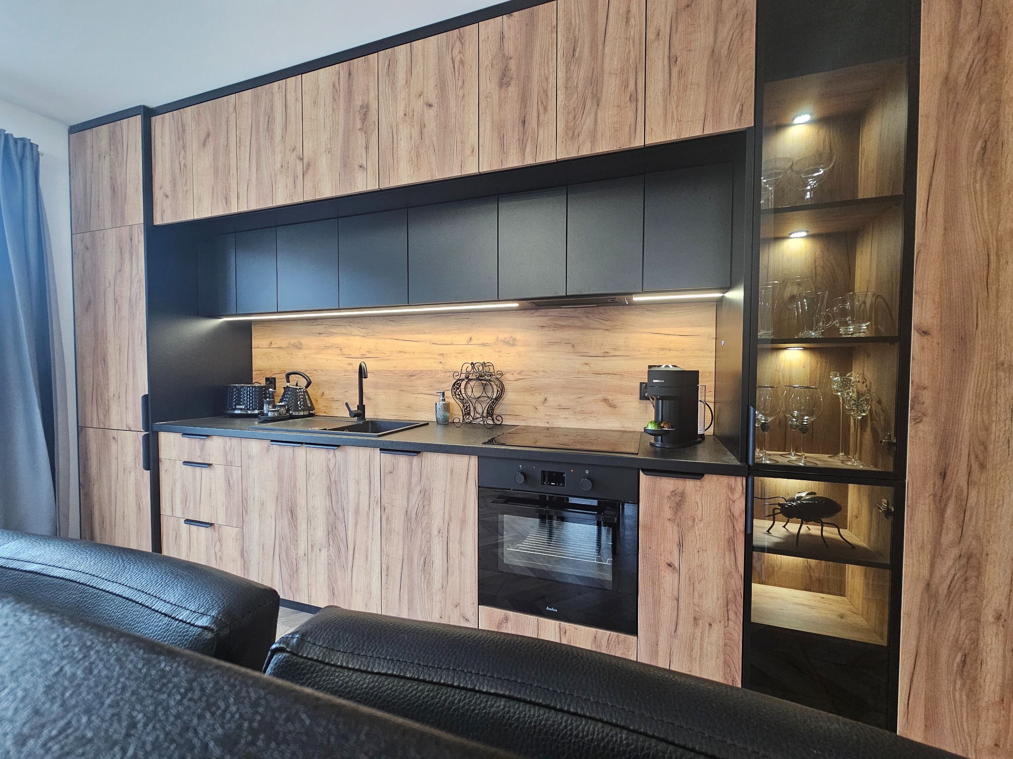 Nowoczesna kuchnia z drewnopodobnymi frontami szafek górnych i dolnych, czarnymi szafkami wiszącymi i blatem, oświetleniem LED pod szafkami, przeszkloną witryną z dekoracyjnym owadem i szkłem...