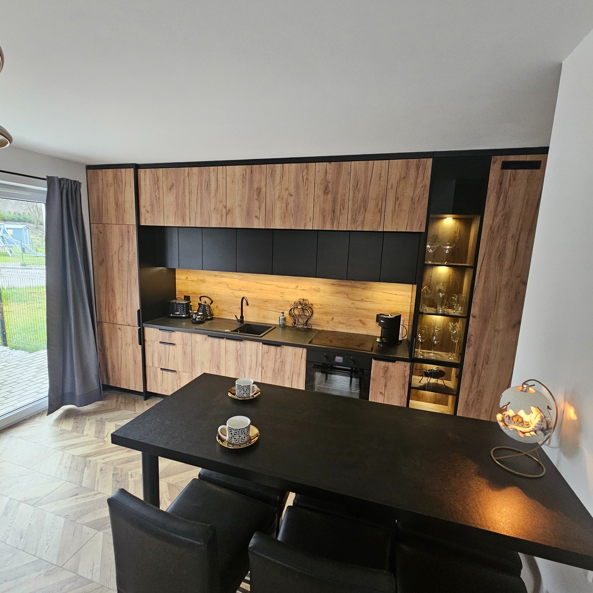 Nowoczesna kuchnia z drewnianymi frontami szafek, czarnymi elementami i oświetleniem LED. Na czarnym stole stoją dwie filiżanki z geometrycznym wzorem, a obok lampka dekoracyjna. Widoczne...