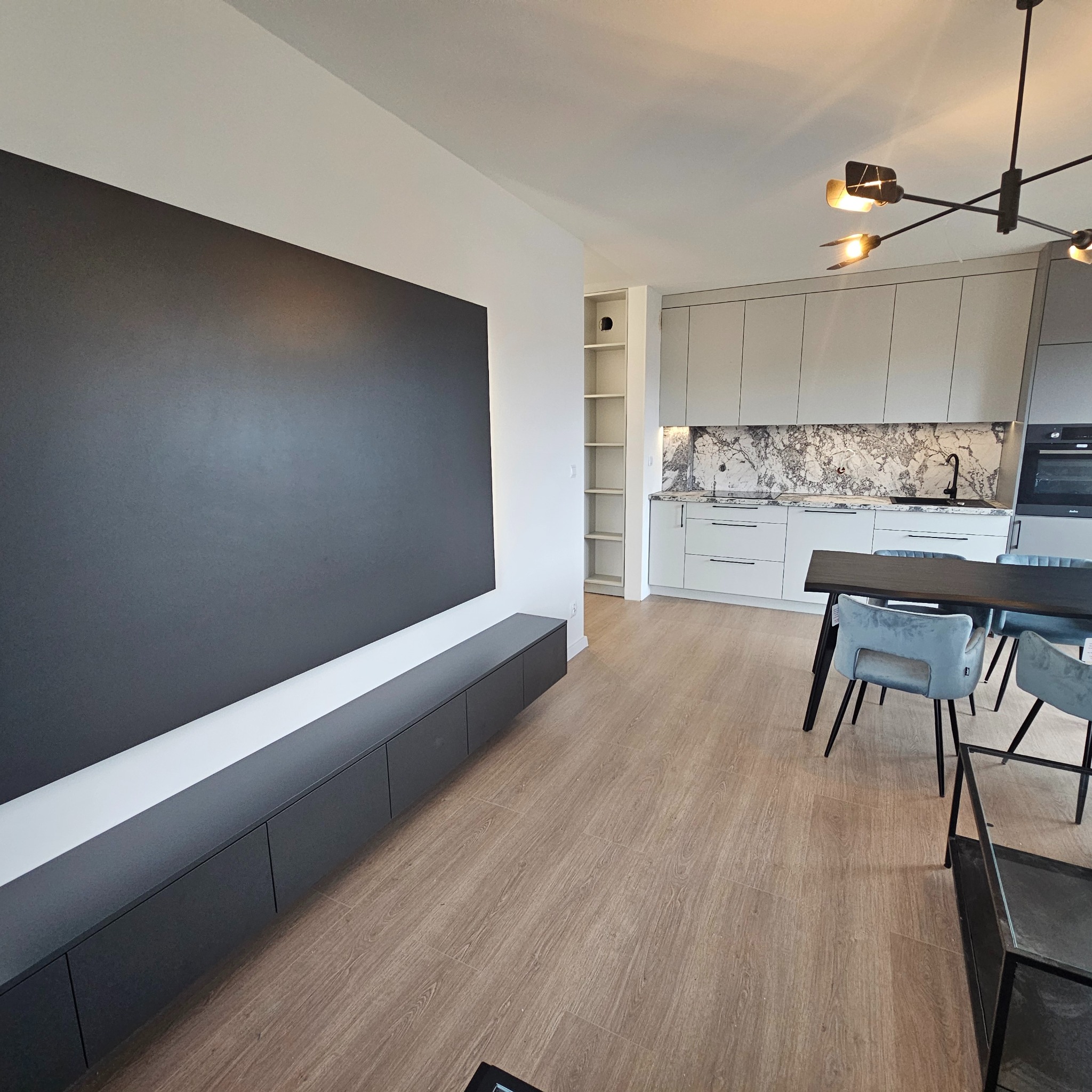 Nowoczesna kuchnia z zabudową w odcieniach szarości, marmurowym backsplashiem i czarną armaturą, widok z salonu z szarą szafką RTV i drewnianą podłogą.