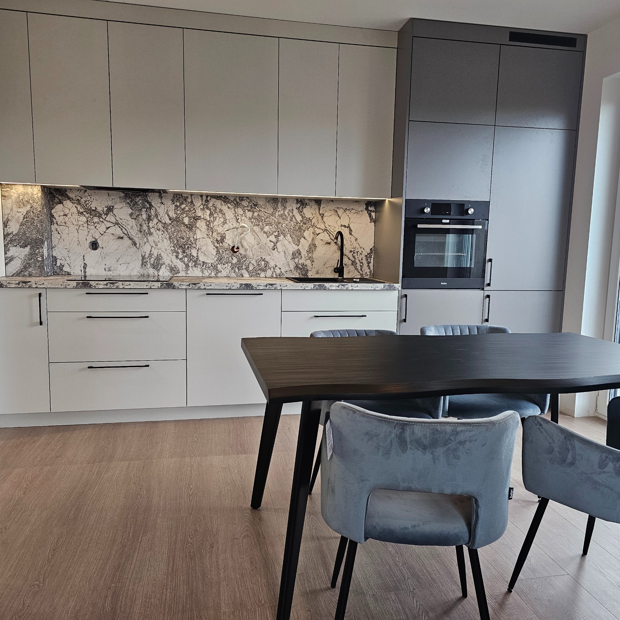 Nowoczesna kuchnia z szarymi szafkami, blatem z motywem marmuru i czarną armaturą, uzupełniona ciemnym drewnianym stołem i szarymi krzesłami w stylu skandynawskim.
