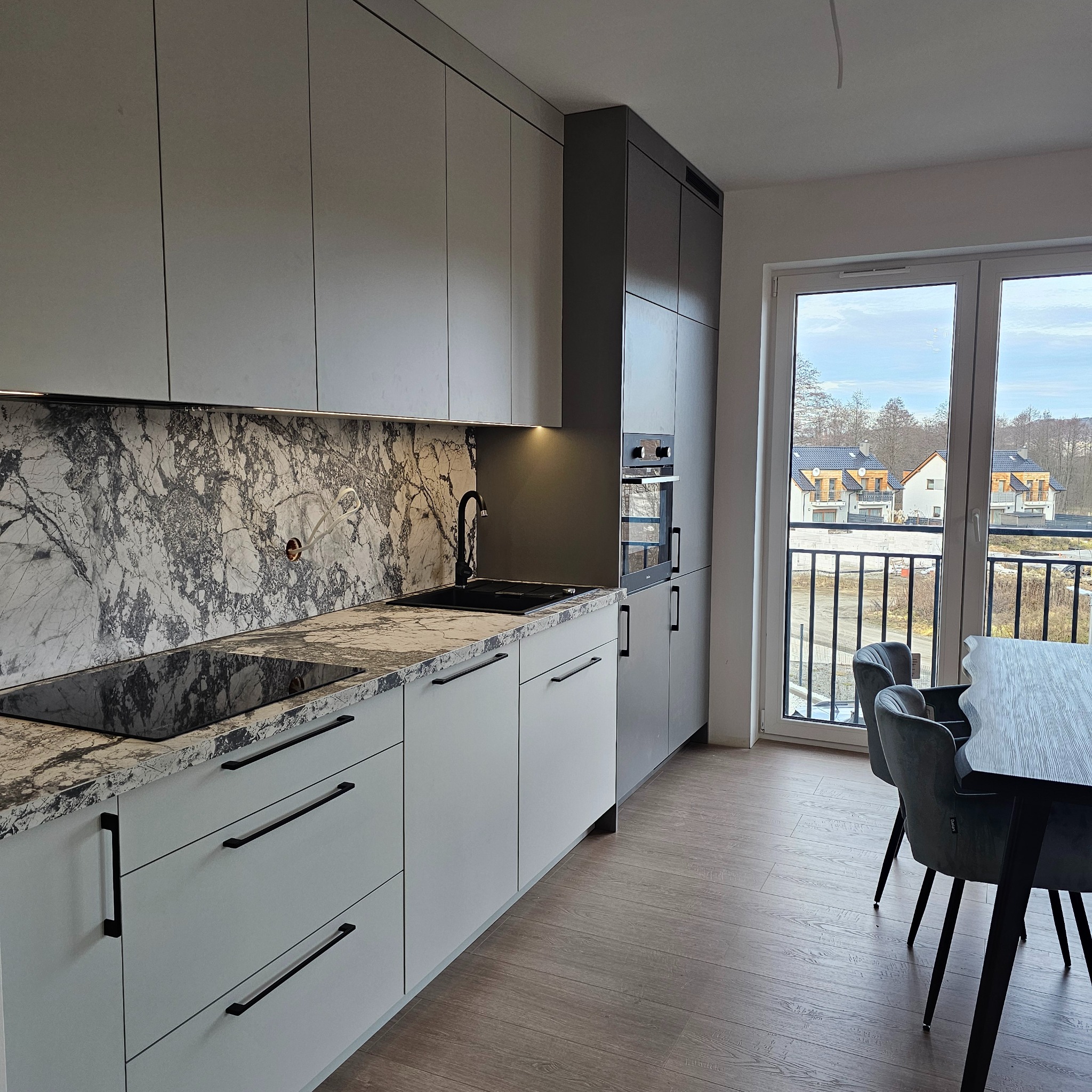 Nowoczesna kuchnia z białymi frontami szafek i blatem z szarego marmuru, czarna armatura i płyta grzewcza, widok na osiedle domów jednorodzinnych przez okno.