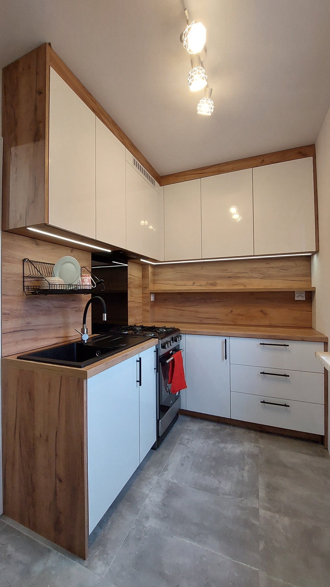 Nowoczesna kuchnia z białymi, lakierowanymi szafkami i drewnopodobnymi akcentami, czarny zlew, czarny kran, oświetlenie LED pod szafkami wiszącymi i designerskie lampy sufitowe.