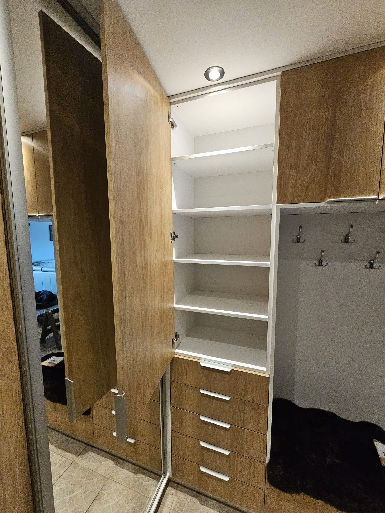 Otwarta szafa wnękowa z przesuwnymi drzwiami lustrzanymi, drewnopodobne fronty, białe półki i szuflady z uchwytami, w tle wieszaki na ubrania i czarny dywanik.