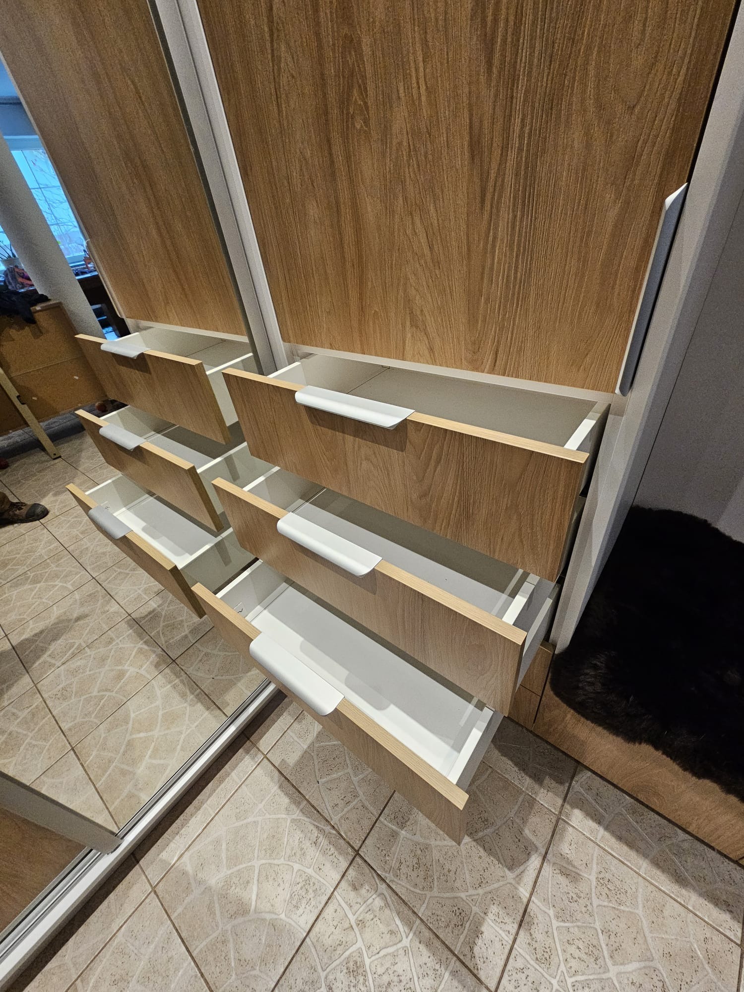 Otwarta szafa przesuwna z drewnopodobnymi frontami i białymi uchwytami szuflad, widoczne wnętrze szuflad i odbicie w lustrzanych drzwiach.