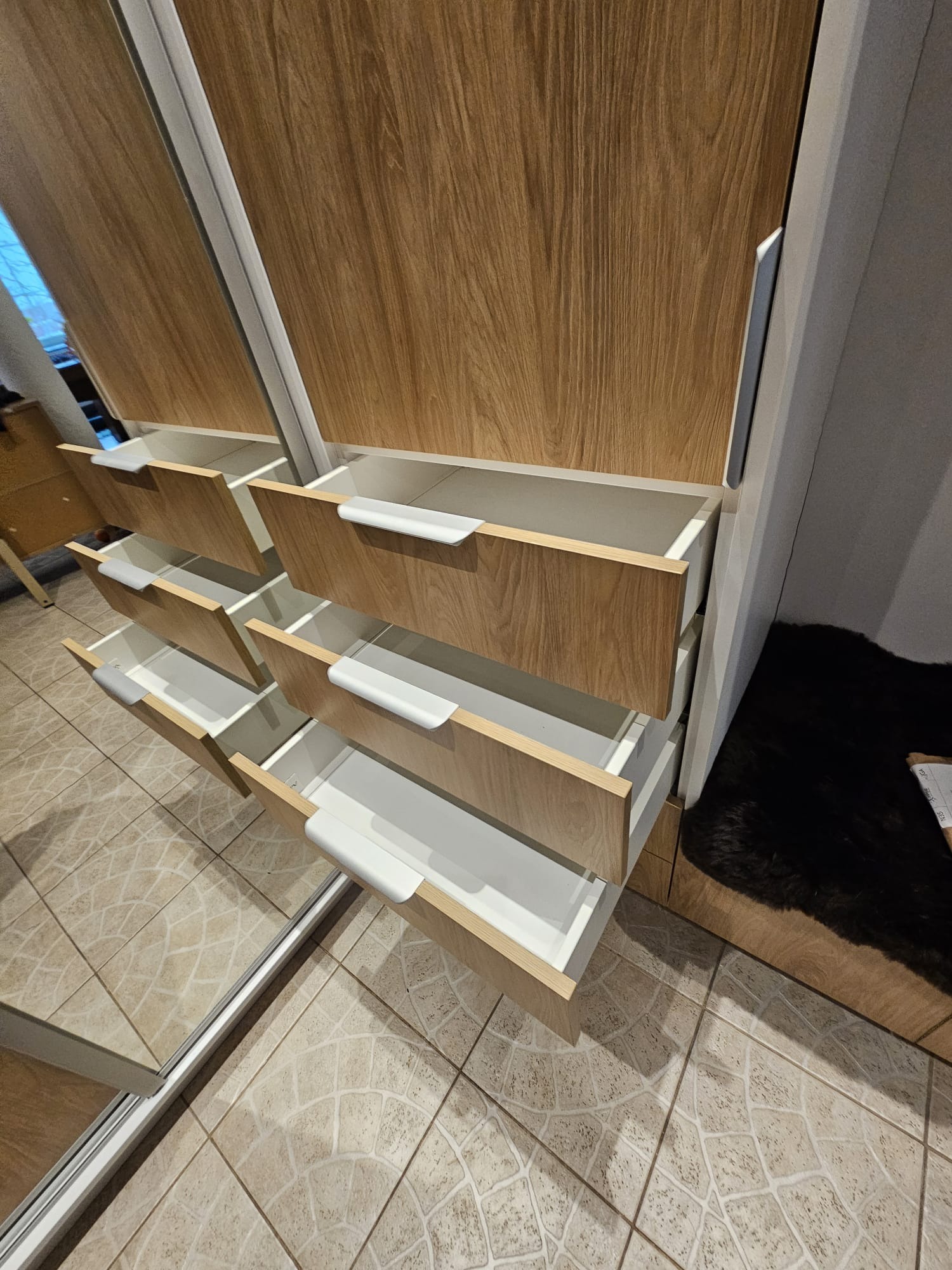 Wnętrze szafy wnękowej z lustrzanymi drzwiami przesuwnymi i otwartymi szufladami z białymi uchwytami, wykończone drewnopodobnym laminatem.