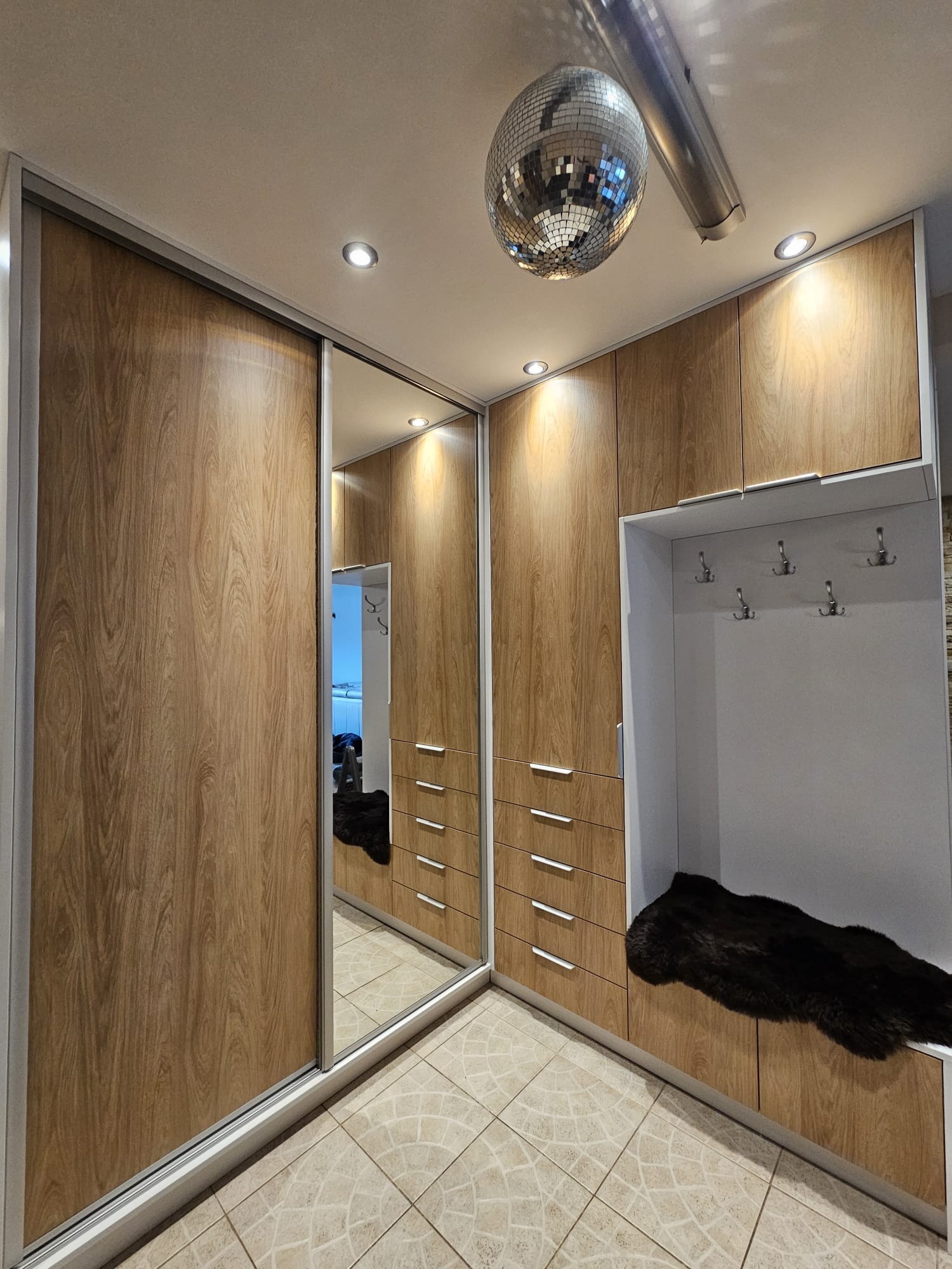 Szafa wnękowa z przesuwnymi drzwiami lustrzanymi i drewnopodobnym wykończeniem, z wbudowanym wieszakiem na ubrania i puszystą narzutą, oświetlona halogenami i dyskotekową kulą.