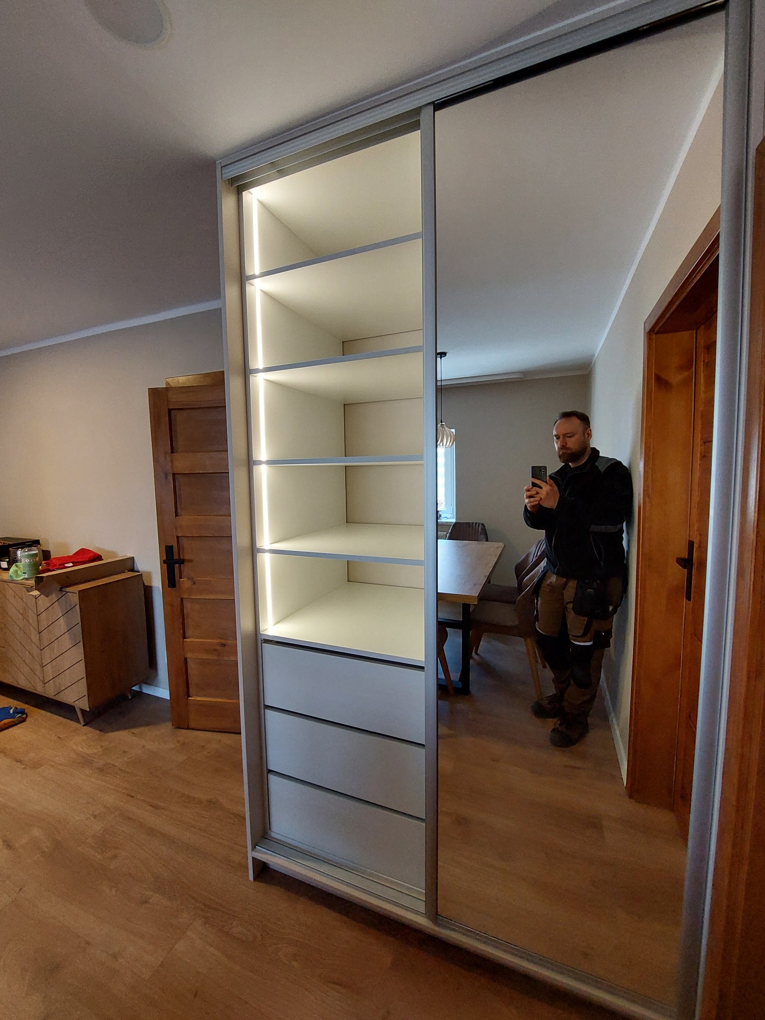 Wnętrze pokoju z nową szafą wnękową z przesuwnymi drzwiami lustrzanymi i oświetleniem LED na półkach, widoczne odbicie mężczyzny robiącego zdjęcie telefonem.
