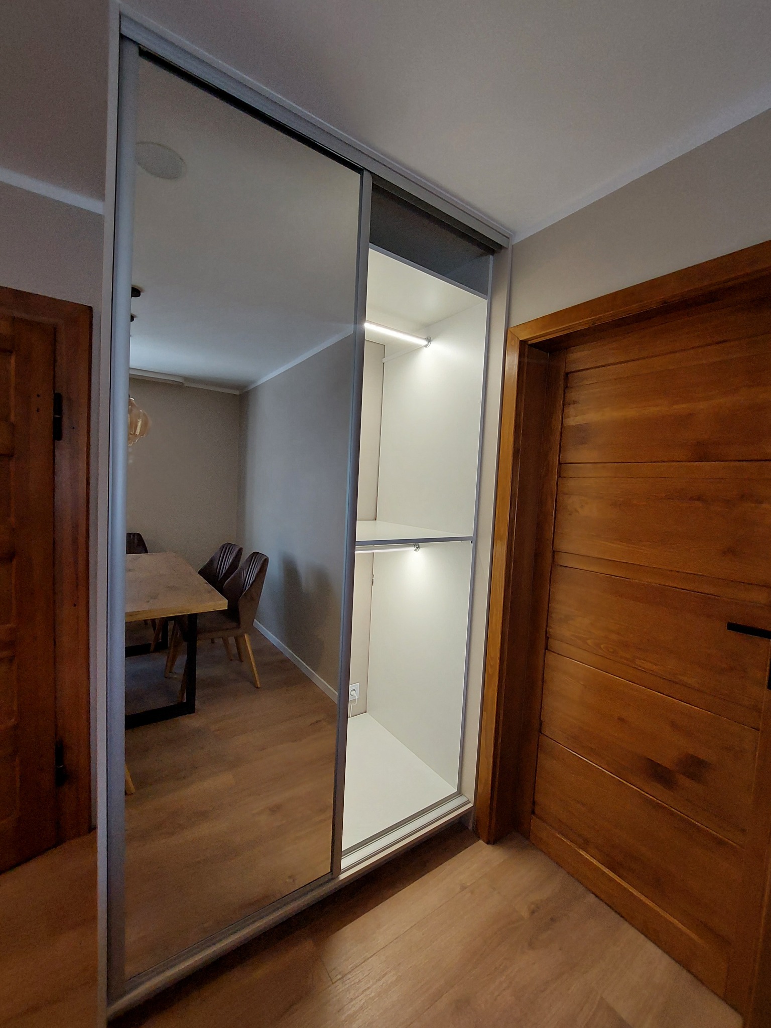 Nowoczesna szafa wnękowa z przesuwnymi drzwiami lustrzanymi i oświetleniem LED, widok z boku na tle drewnianych drzwi i fragmentu jadalni.