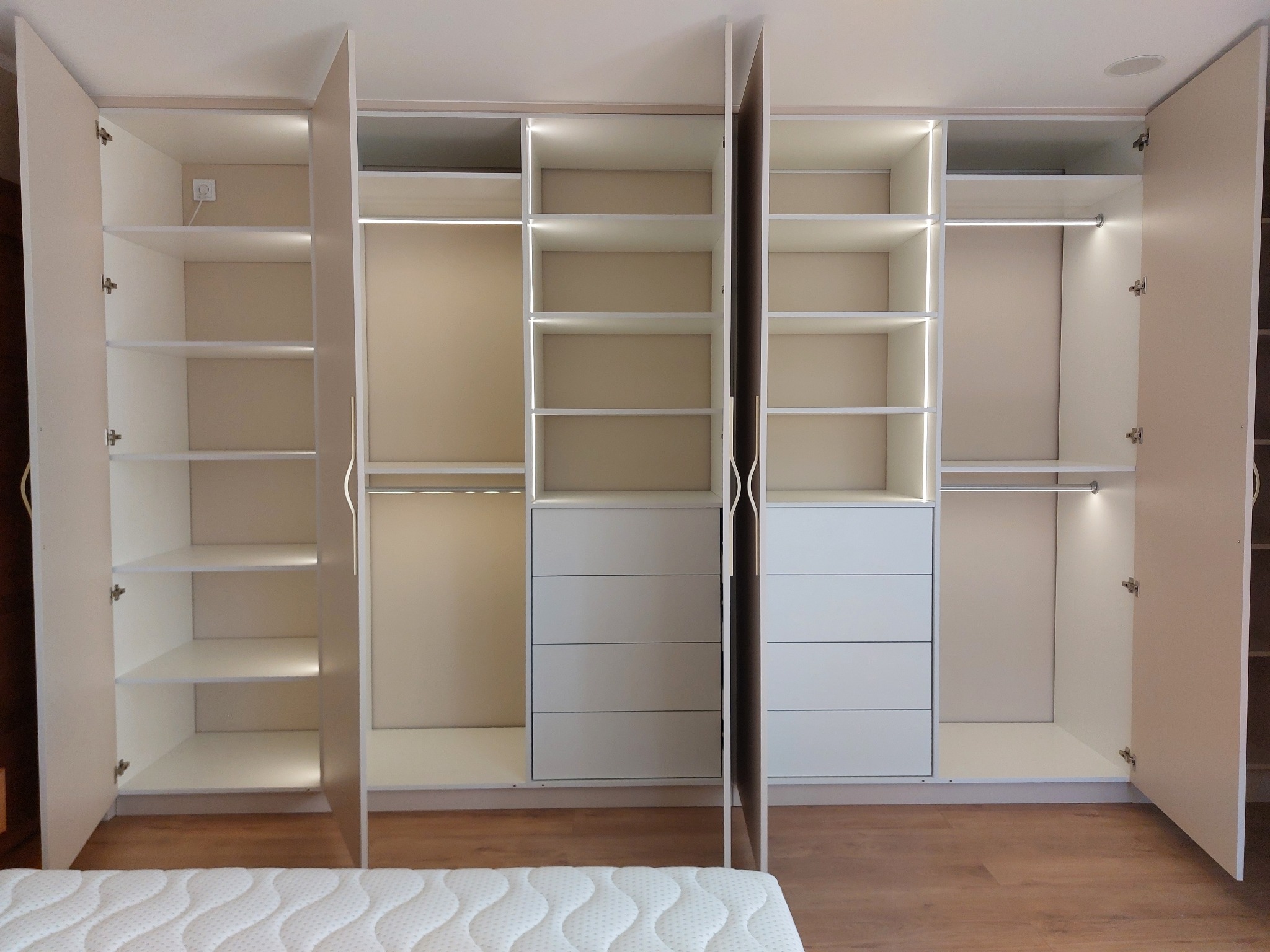 Wnętrze szafy wnękowej z oświetleniem LED, widoczne półki, szuflady i drążek na ubrania, z otwartymi drzwiami po bokach, na podłodze panele, w tle fragment materaca.