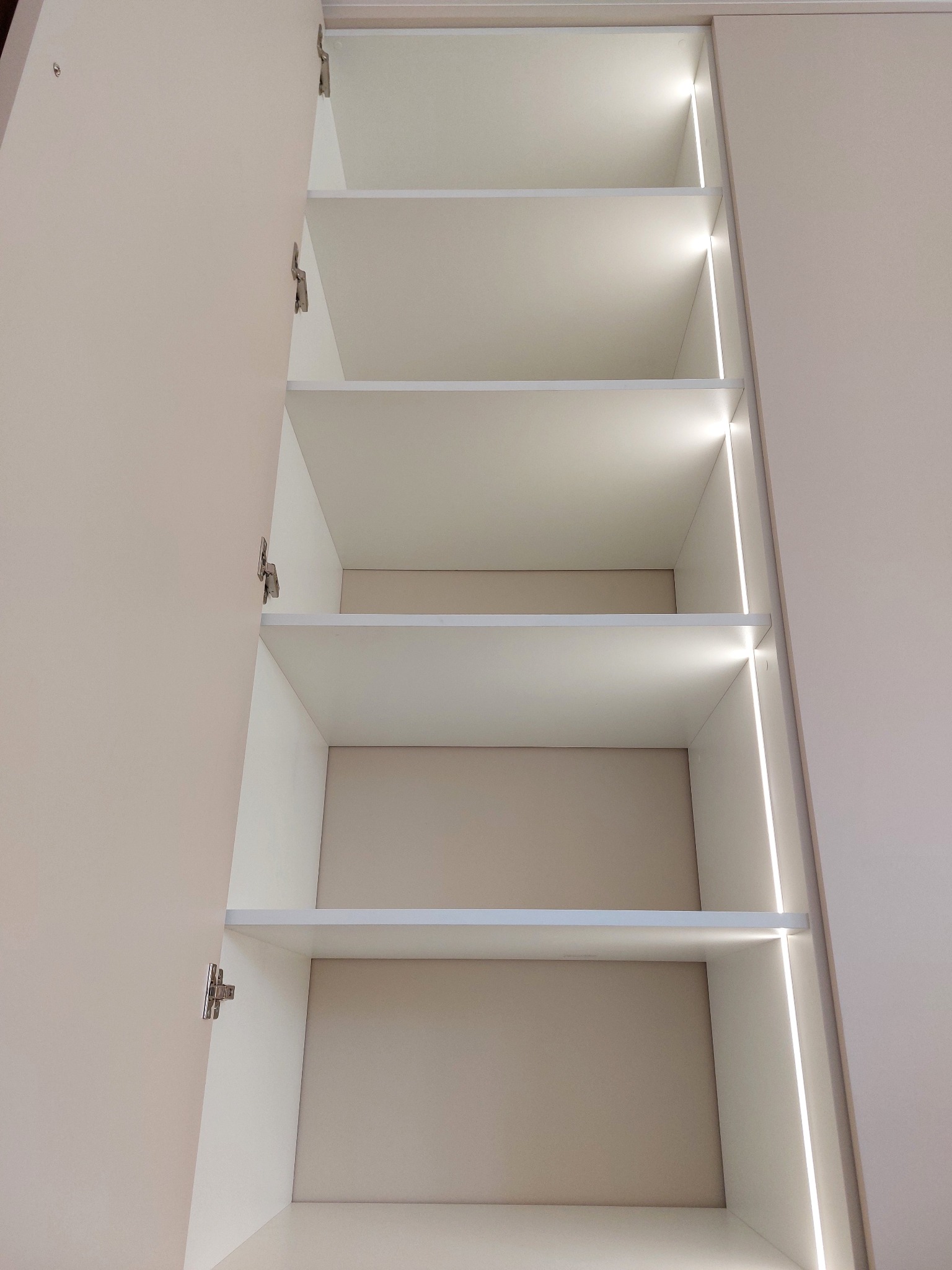 Wnętrze szafy wnękowej z otwartymi drzwiami, wyposażonej w sześć półek i pionowe oświetlenie LED wzdłuż krawędzi, widok z dołu.