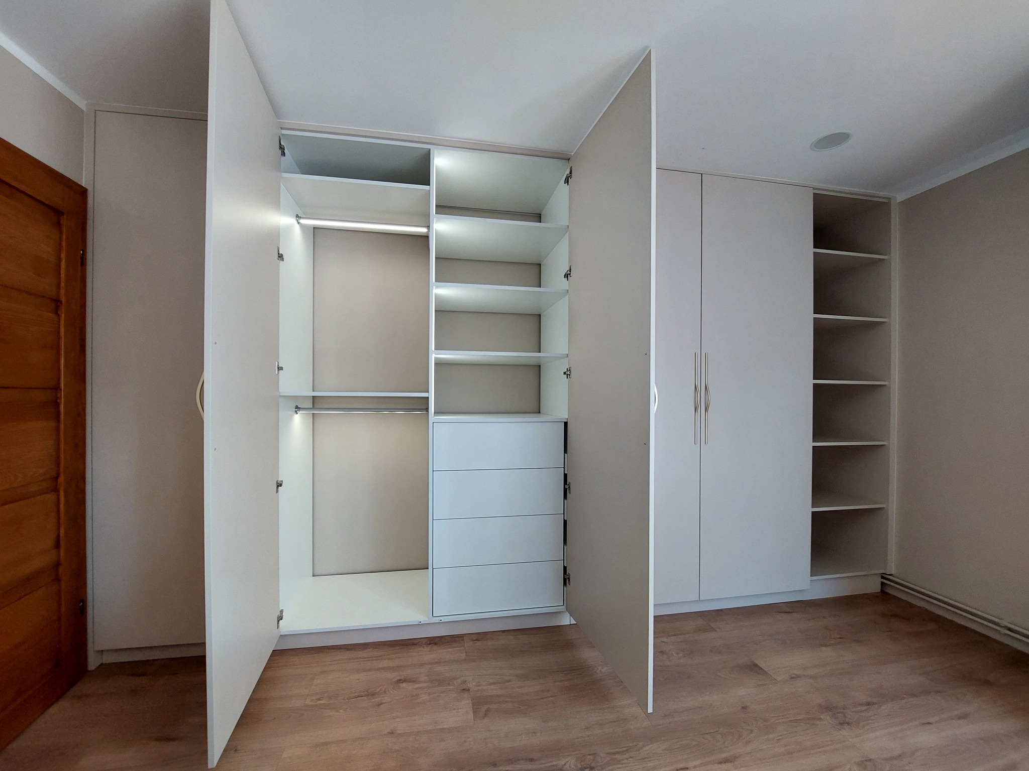 Jasnoszara szafa wnękowa z otwartymi drzwiami, podświetlana od wewnątrz, z półkami, drążkami i czterema szufladami, na podłodze panele imitujące drewno.