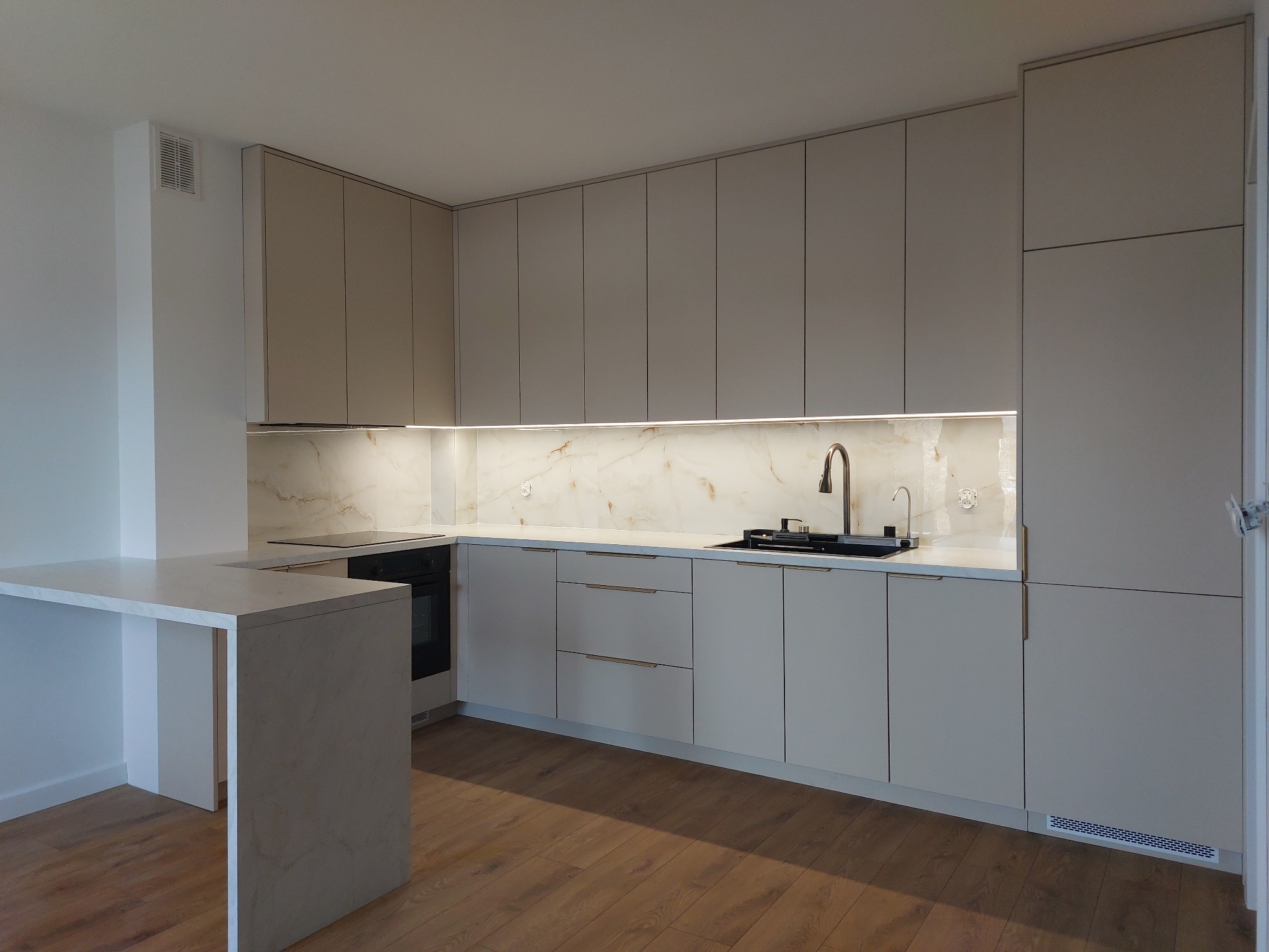 Nowoczesna kuchnia z jasnymi, gładkimi frontami szafek, oświetleniem LED pod szafkami wiszącymi i blatem imitującym marmur, widoczny fragment blatu z barkiem