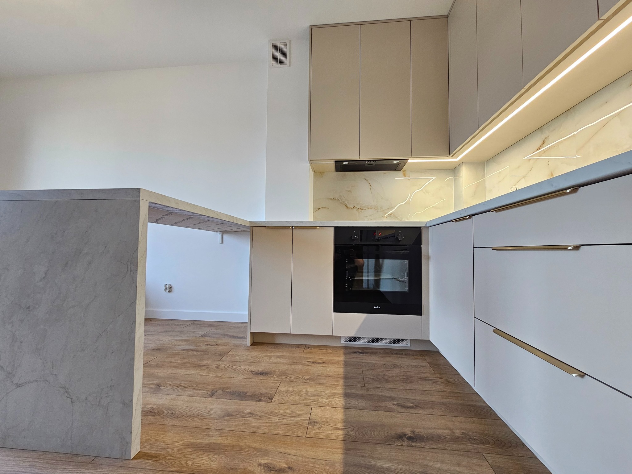 Nowoczesna kuchnia z beżowymi szafkami górnymi i białymi dolnymi, oświetleniem LED podszafkowym i blatem imitującym marmur, widziana z niskiego kąta.