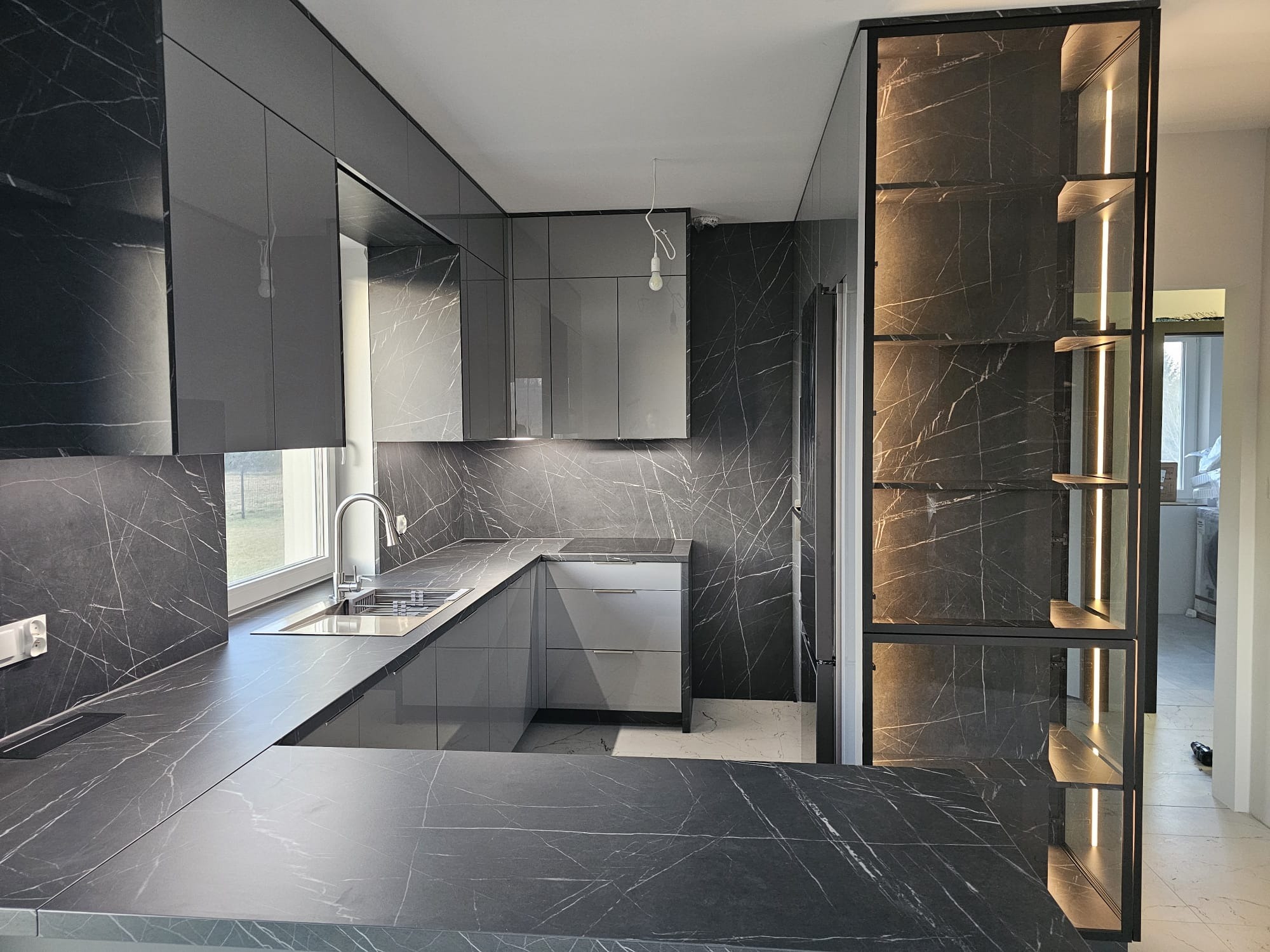 Nowoczesna, szara kuchnia z blatem imitującym marmur, oświetleniem LED i otwartą półką z podświetleniem. Widok na okno i fragment podłogi.