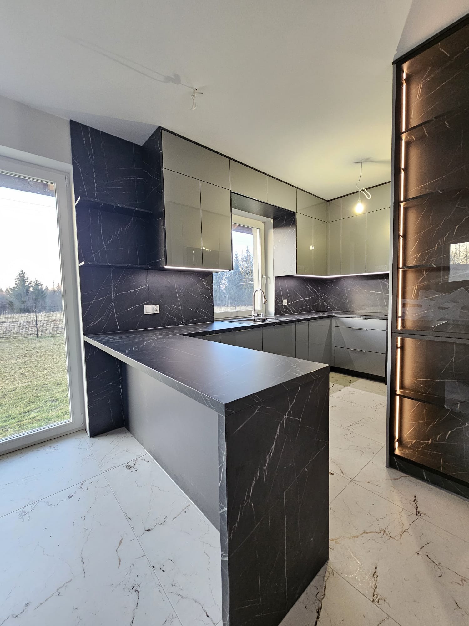 Nowoczesna kuchnia z szarymi szafkami i blatem z czarnego marmuru z białymi żyłami. Wyspa kuchenna z blatem z tego samego materiału. Oświetlenie LED pod szafkami.