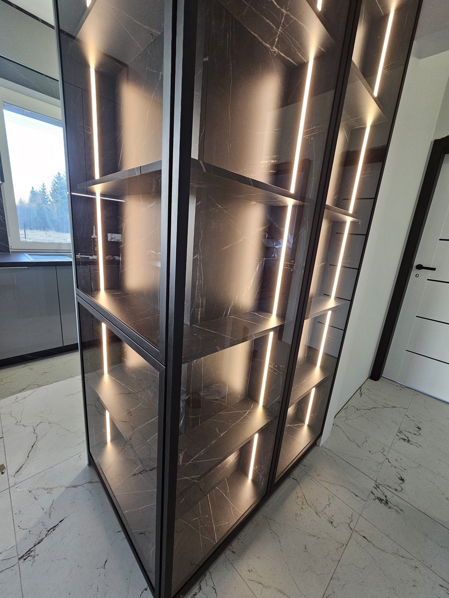 Nowoczesna szafa wnękowa z ciemnego marmuru, podświetlana pionowymi paskami LED, z widocznym fragmentem kuchni i okna w tle. Eleganckie wykończenie wnętrza.
