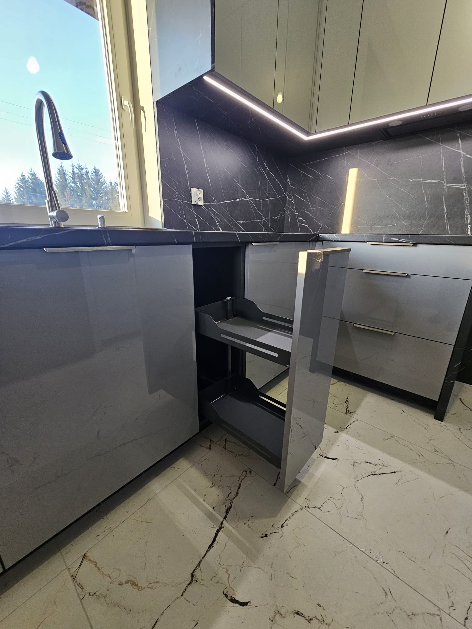 Szara kuchnia z marmurowym blatem i wysuwanym koszem cargo. Nowoczesny design z oświetleniem LED i widokiem na zewnątrz przez okno. Eleganckie wykończenie.