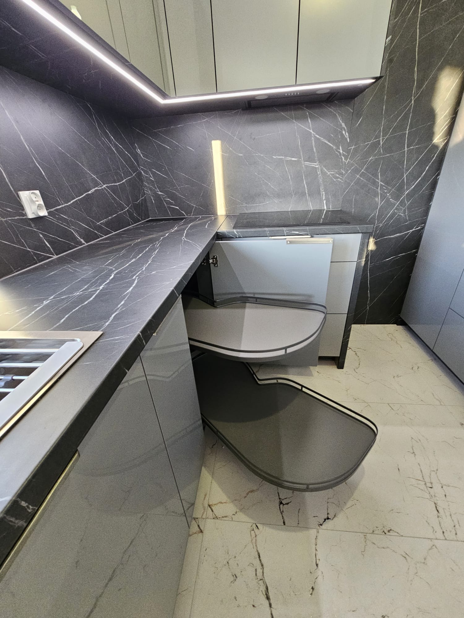 Szara kuchnia z blatem imitującym marmur, z wysuwanymi półkami narożnymi. Nowoczesne oświetlenie LED podkreśla wzór kamienia na ścianie.