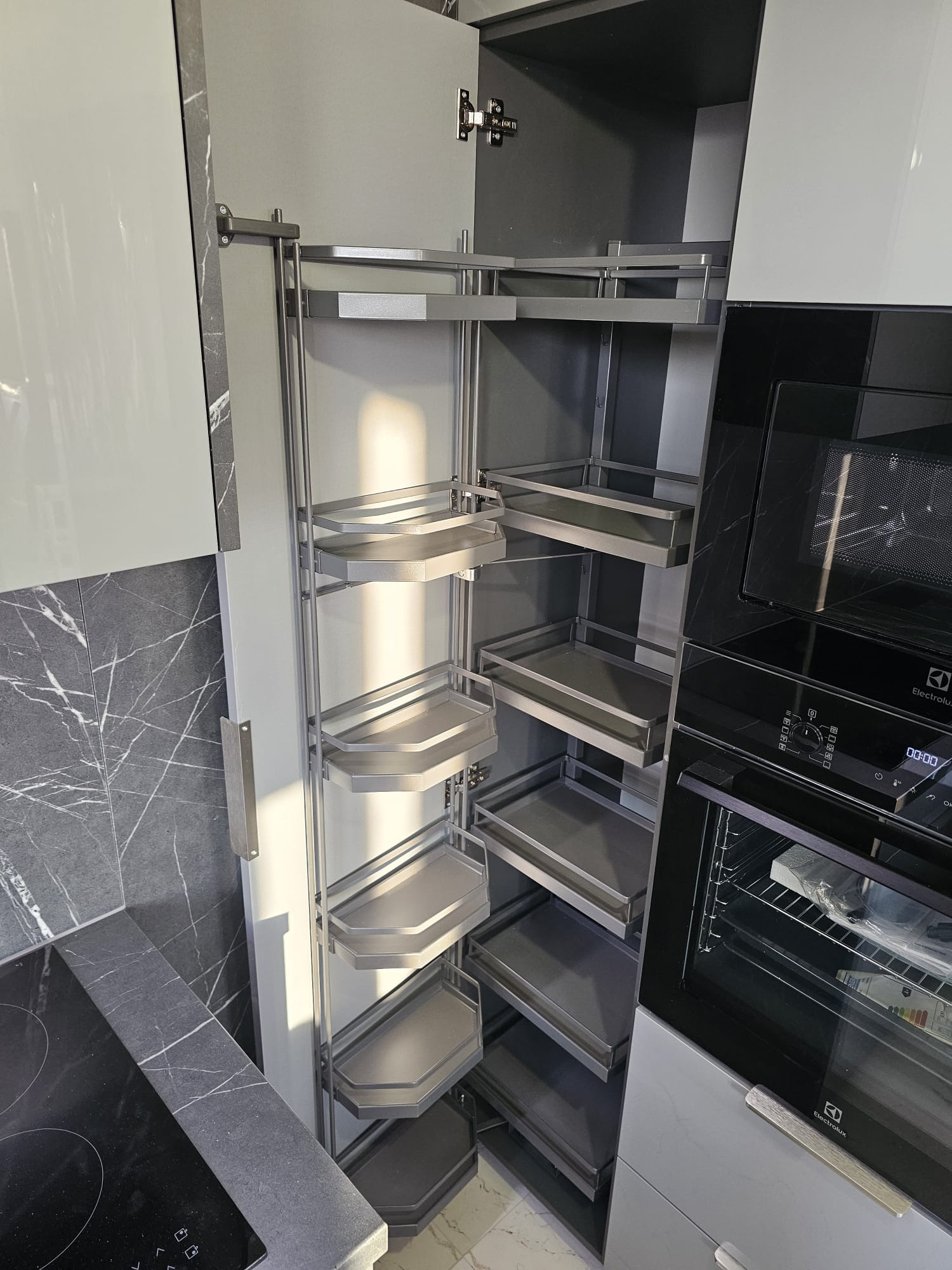 Nowoczesna kuchnia z szarymi frontami i blatem imitującym marmur. Otwarta szafka cargo z półkami obrotowymi, obok piekarnik Electrolux w zabudowie.