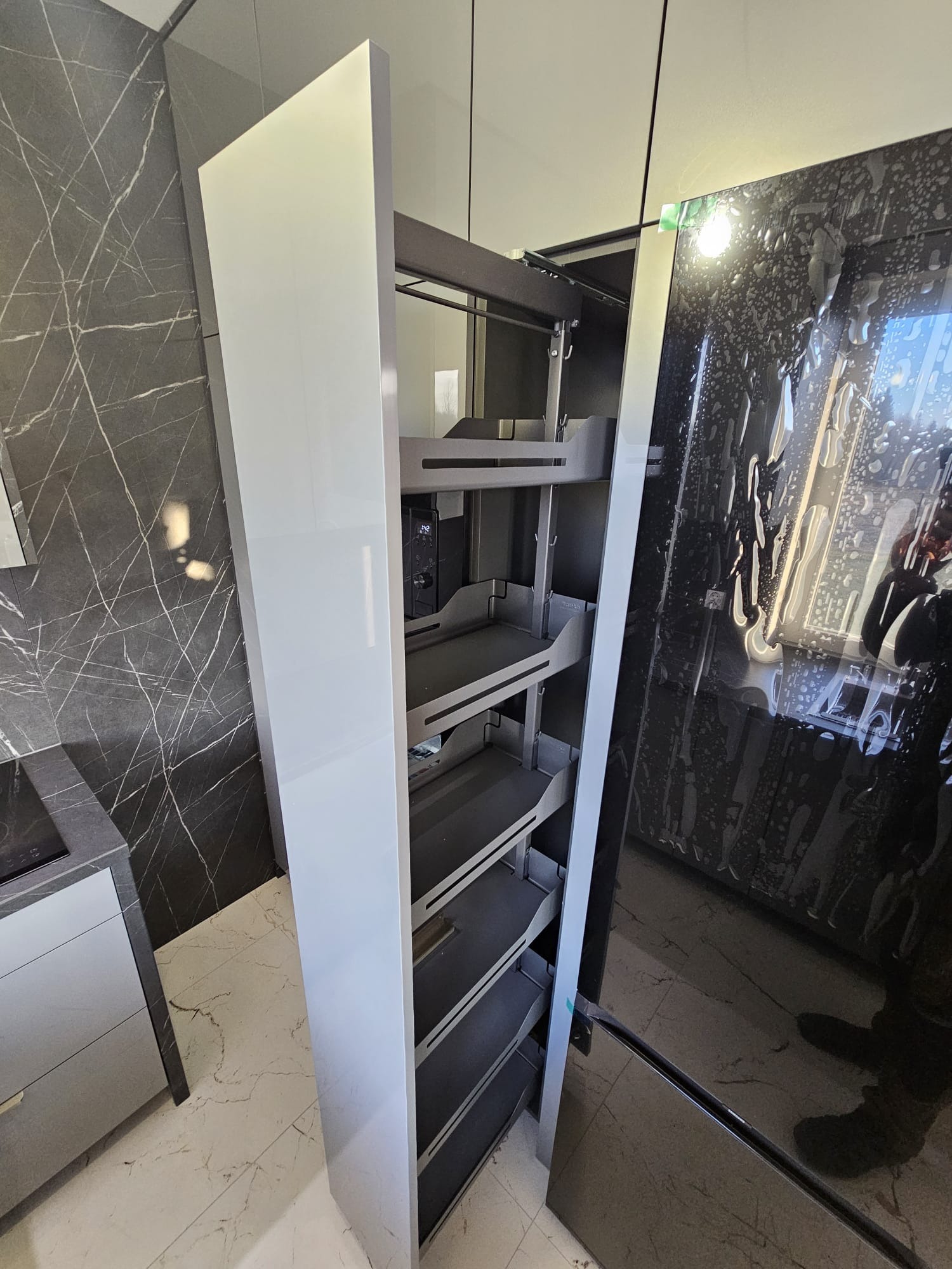 Wysuwana szafka cargo w nowoczesnej kuchni z marmurowym blatem i ścianą. Szafka z szarymi półkami, widoczna na tle błyszczących, czarnych frontów.