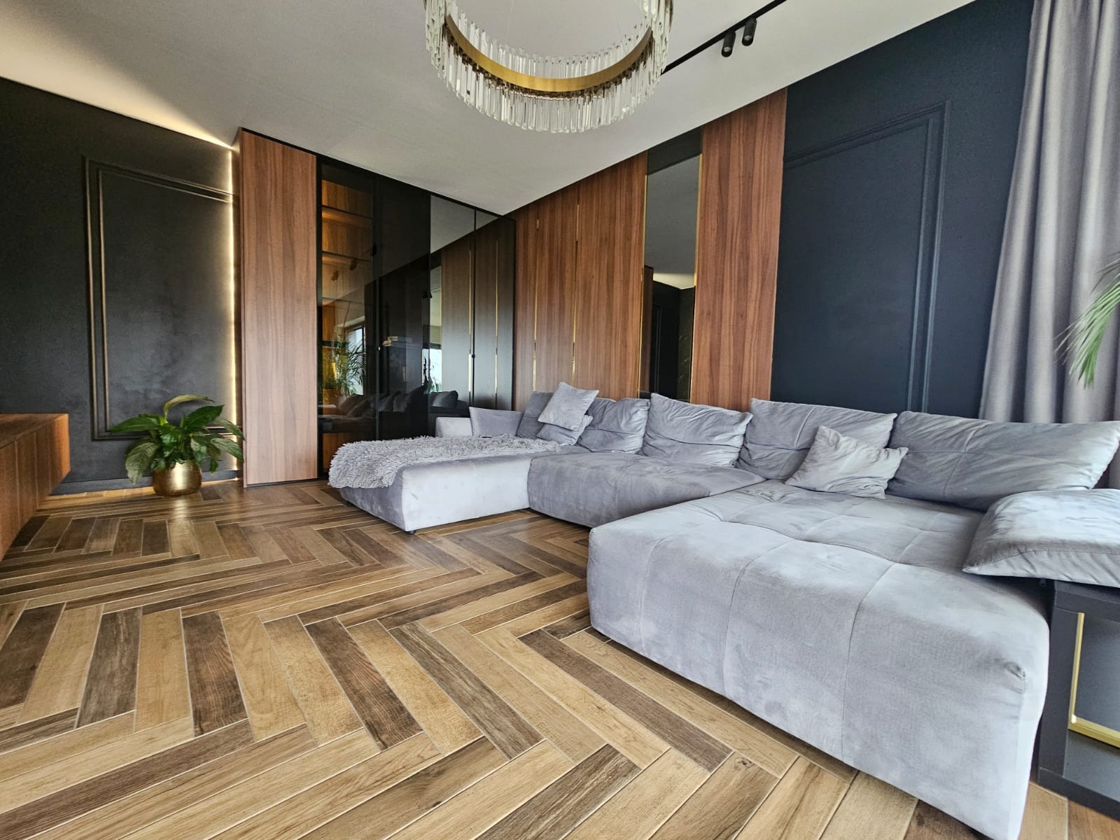 Nowoczesny salon z szarą, modułową sofą narożną, parkietem w jodełkę i ścianami z drewnianymi panelami. Widoczny żyrandol i roślina w złotej doniczce.