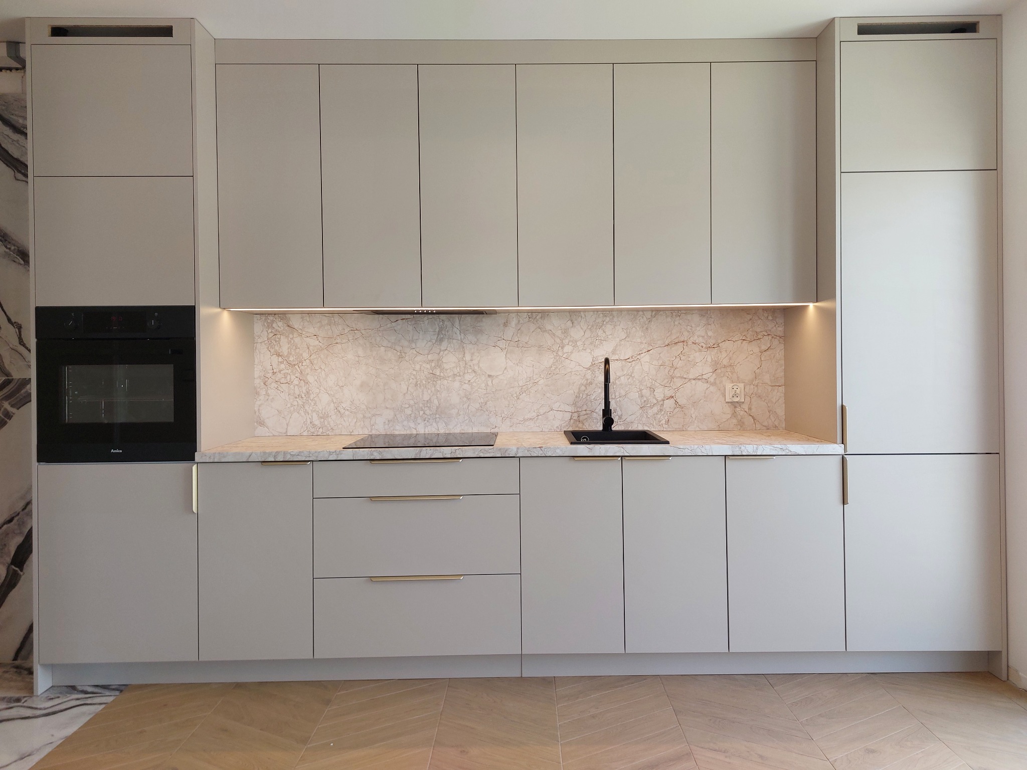 Jasnoszara zabudowa kuchenna z marmurowym blatem i złotymi uchwytami, oświetlona listwą LED. Nowoczesny design z czarną armaturą i sprzętem AGD.