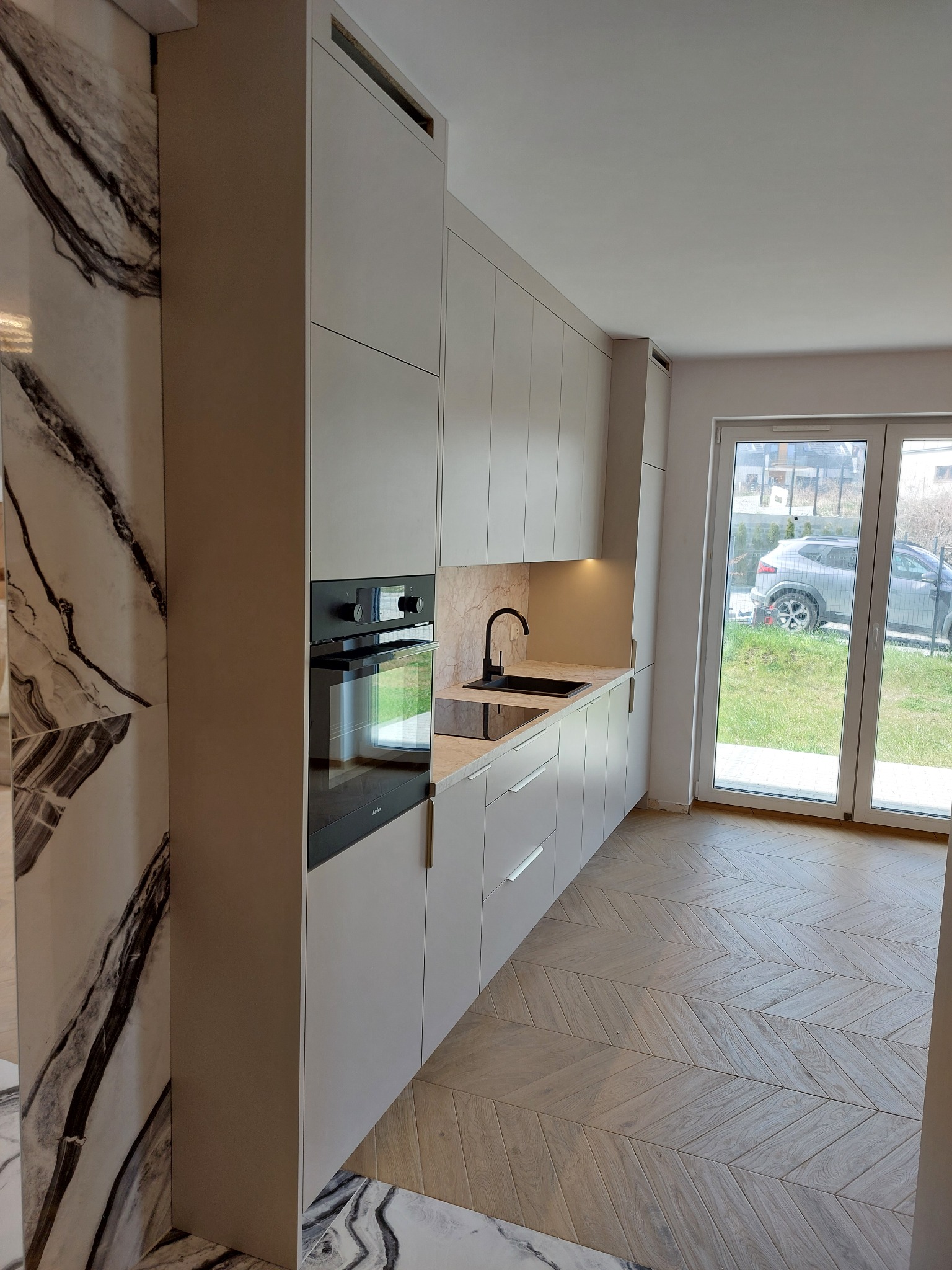 Jasna kuchnia na wymiar z czarną armaturą i piekarnikiem, marmurowy panel boczny, parkiet w jodełkę, okno balkonowe z widokiem na zieleń.