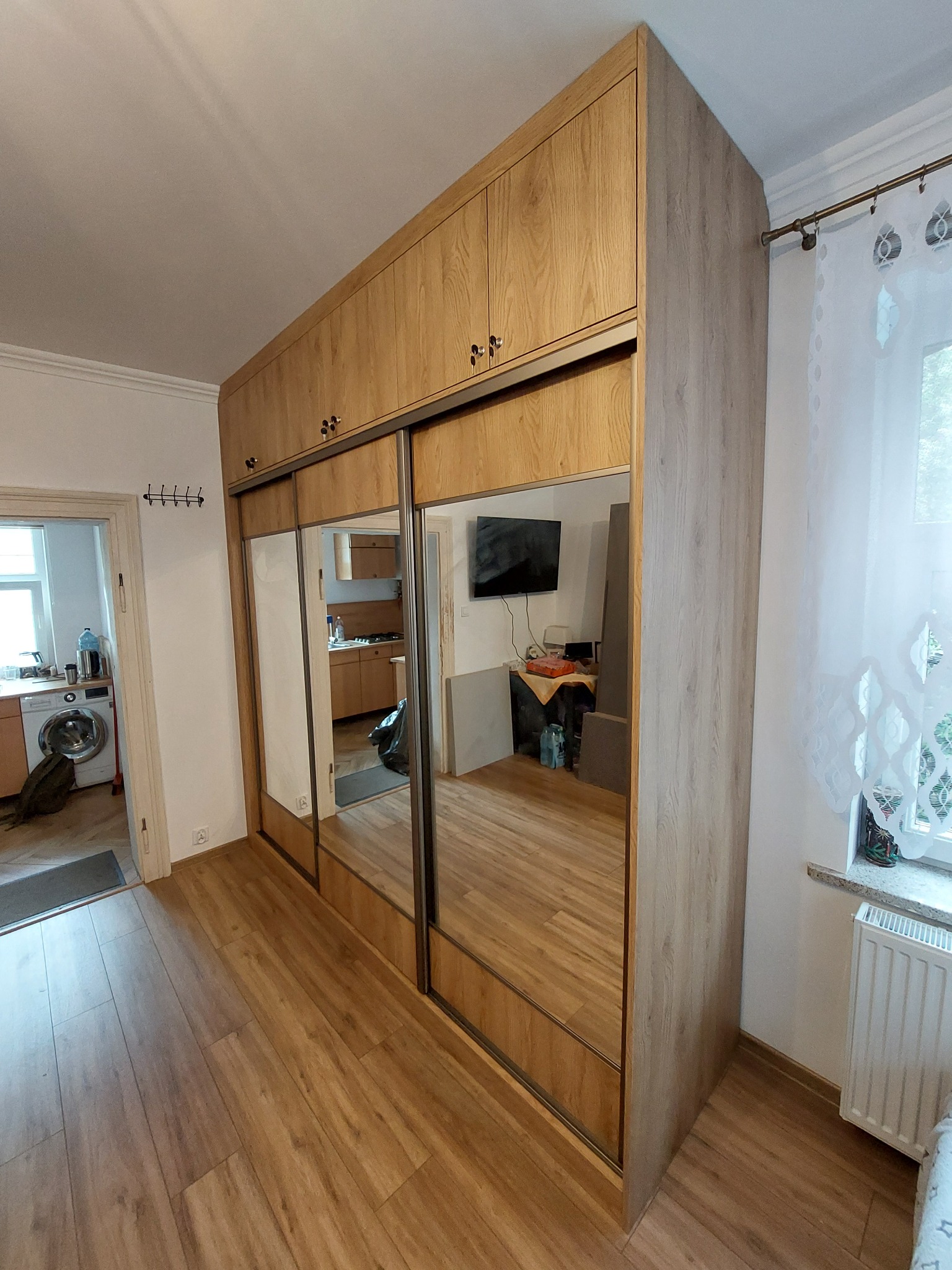 Jasna szafa wnękowa z lustrami, drewnopodobne wykończenie, przesuwne drzwi. Widok na wnętrze mieszkania w odbiciu luster.