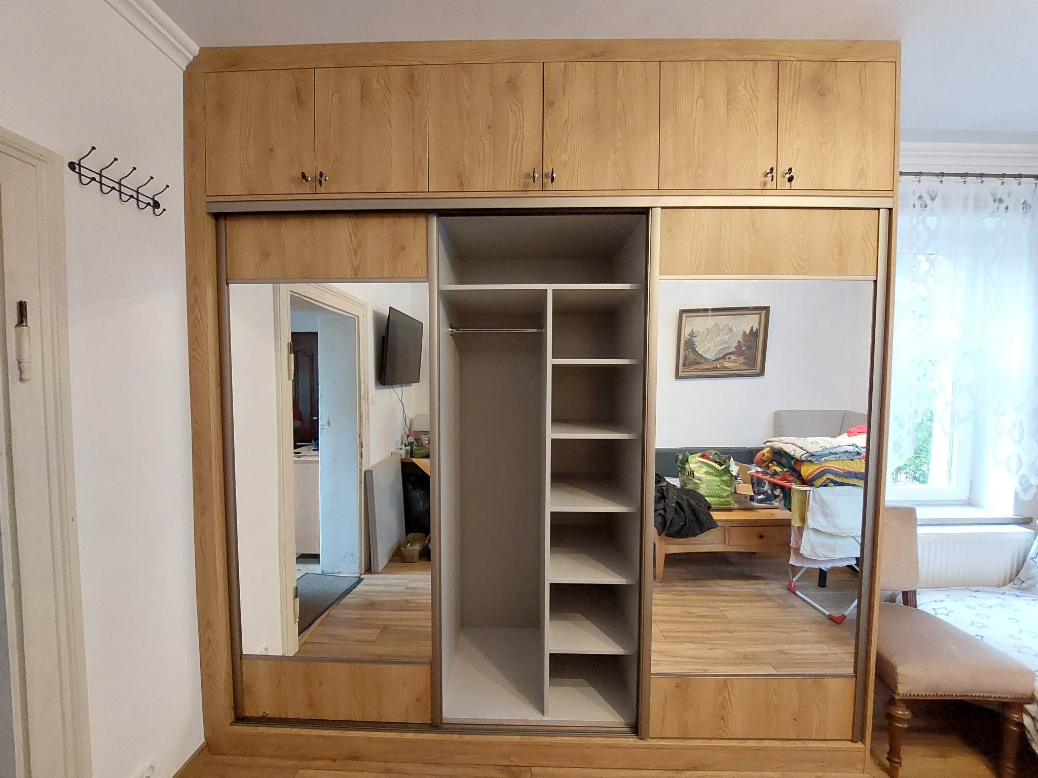 Drewniana szafa wnękowa z przesuwnymi drzwiami lustrzanymi, widok na wnętrze z półkami. Nadstawka z szafkami. Widoczne odbicie pokoju w lustrach.