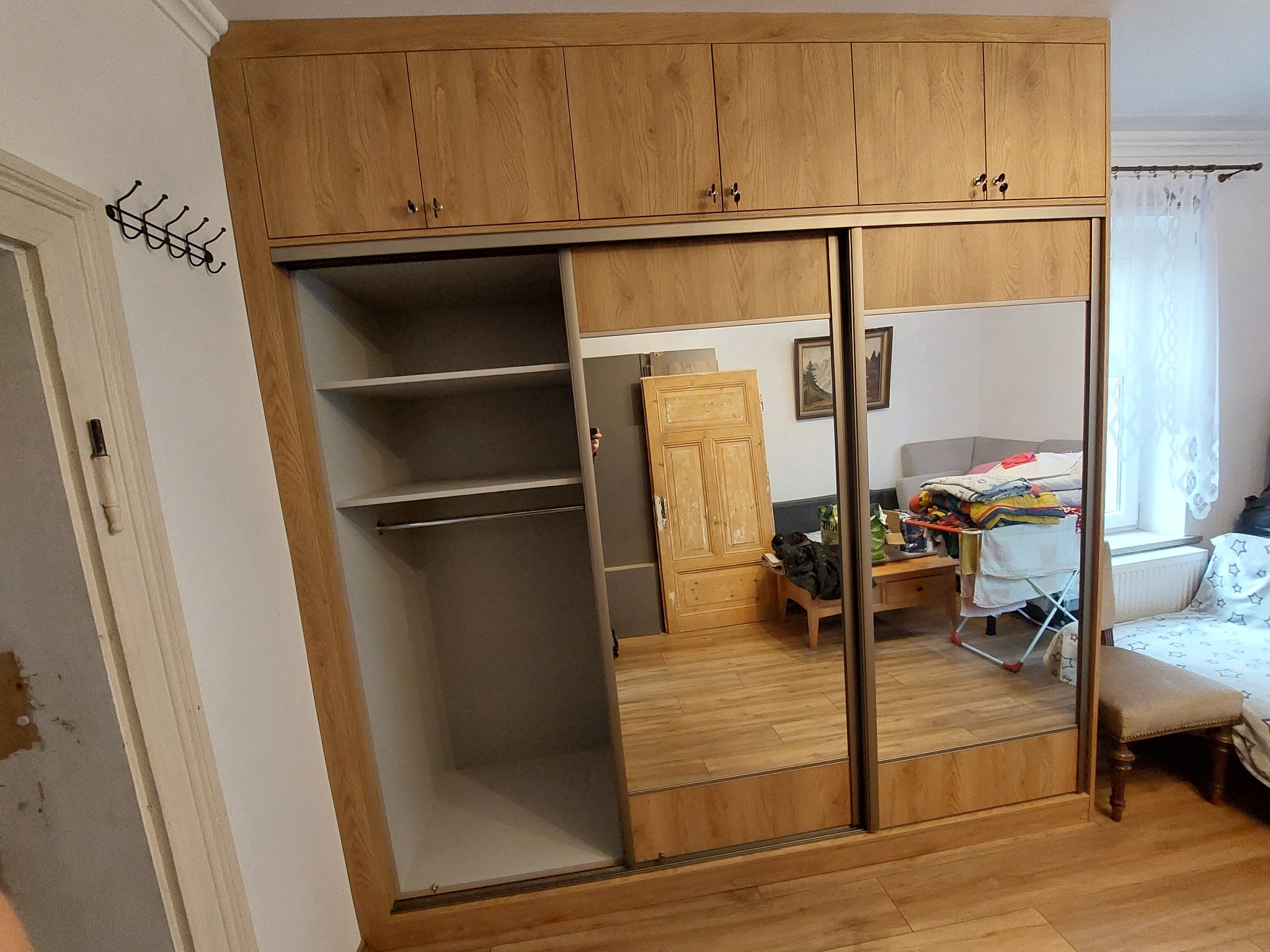 Jasna szafa przesuwna z lustrami, drewnopodobne wykończenie, widoczne półki i drążek na ubrania. W lustrze odbicie pokoju z meblami i oknem.