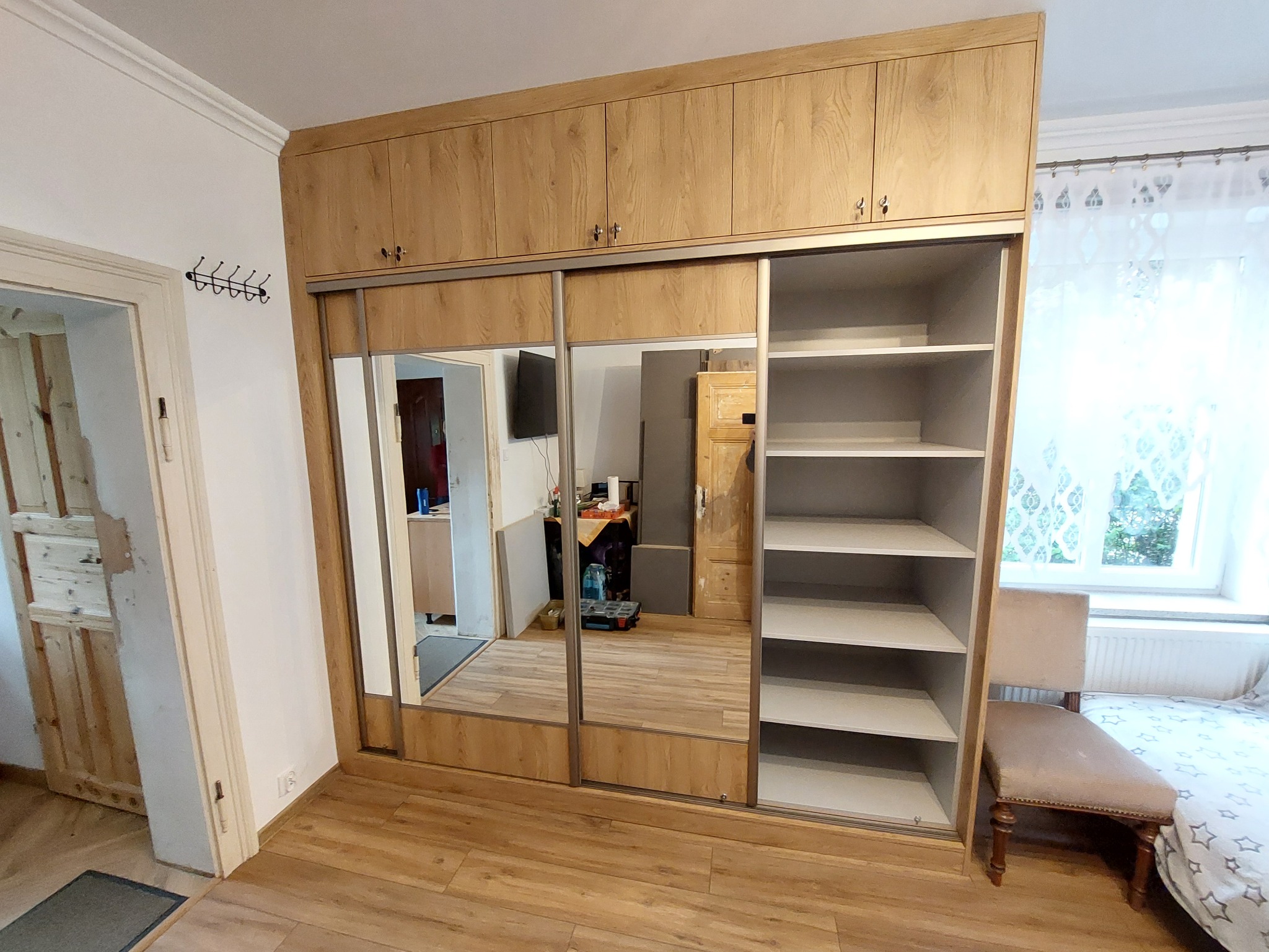 Jasna szafa wnękowa z przesuwnymi drzwiami lustrzanymi, z górnymi szafkami, dopasowana do wnętrza pokoju. Widoczne półki i krzesło obok.